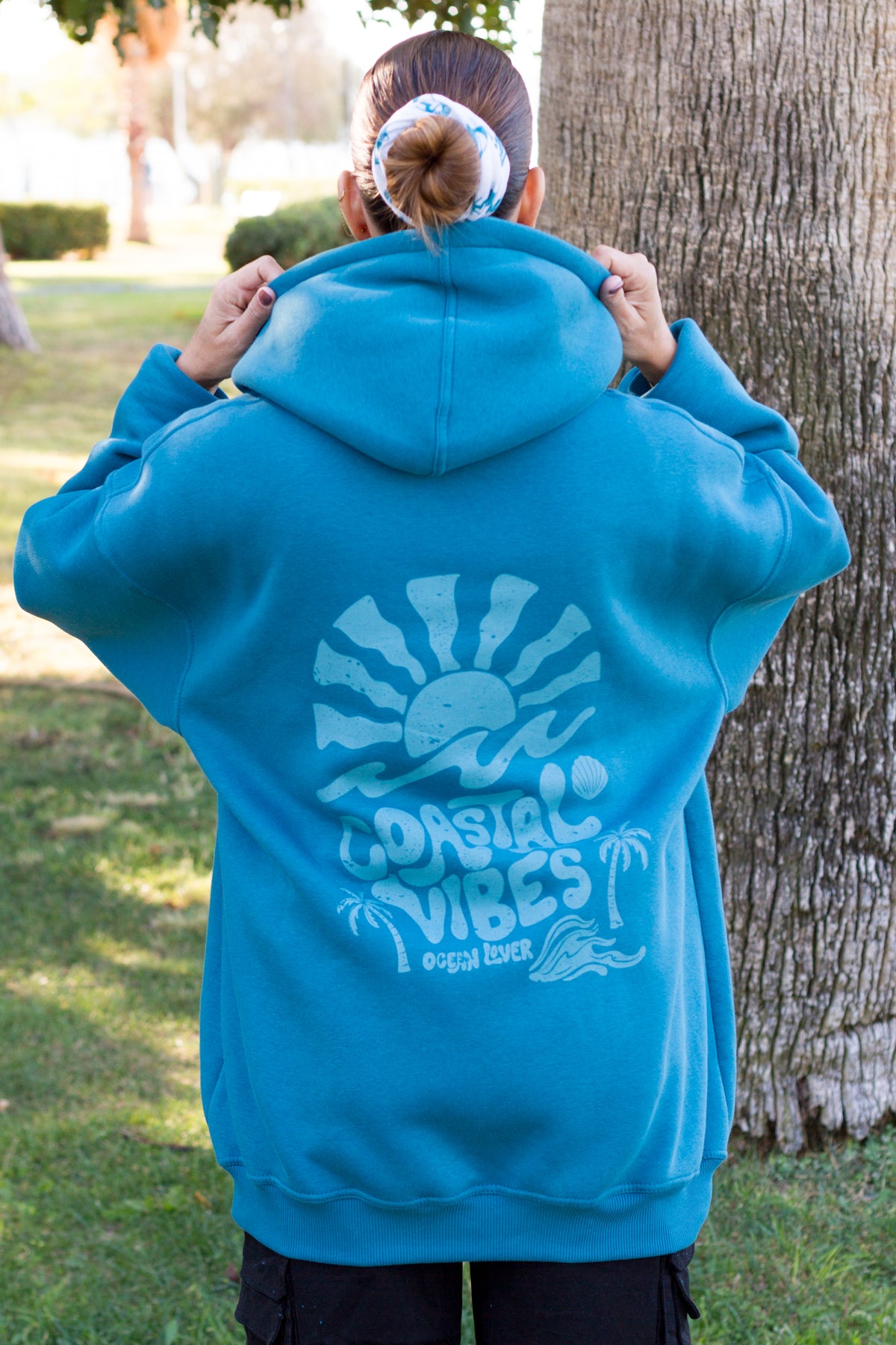 Back view of the petrol TIDE SHIFT hoodie featuring a retro coastal illustration with palm trees and shells.