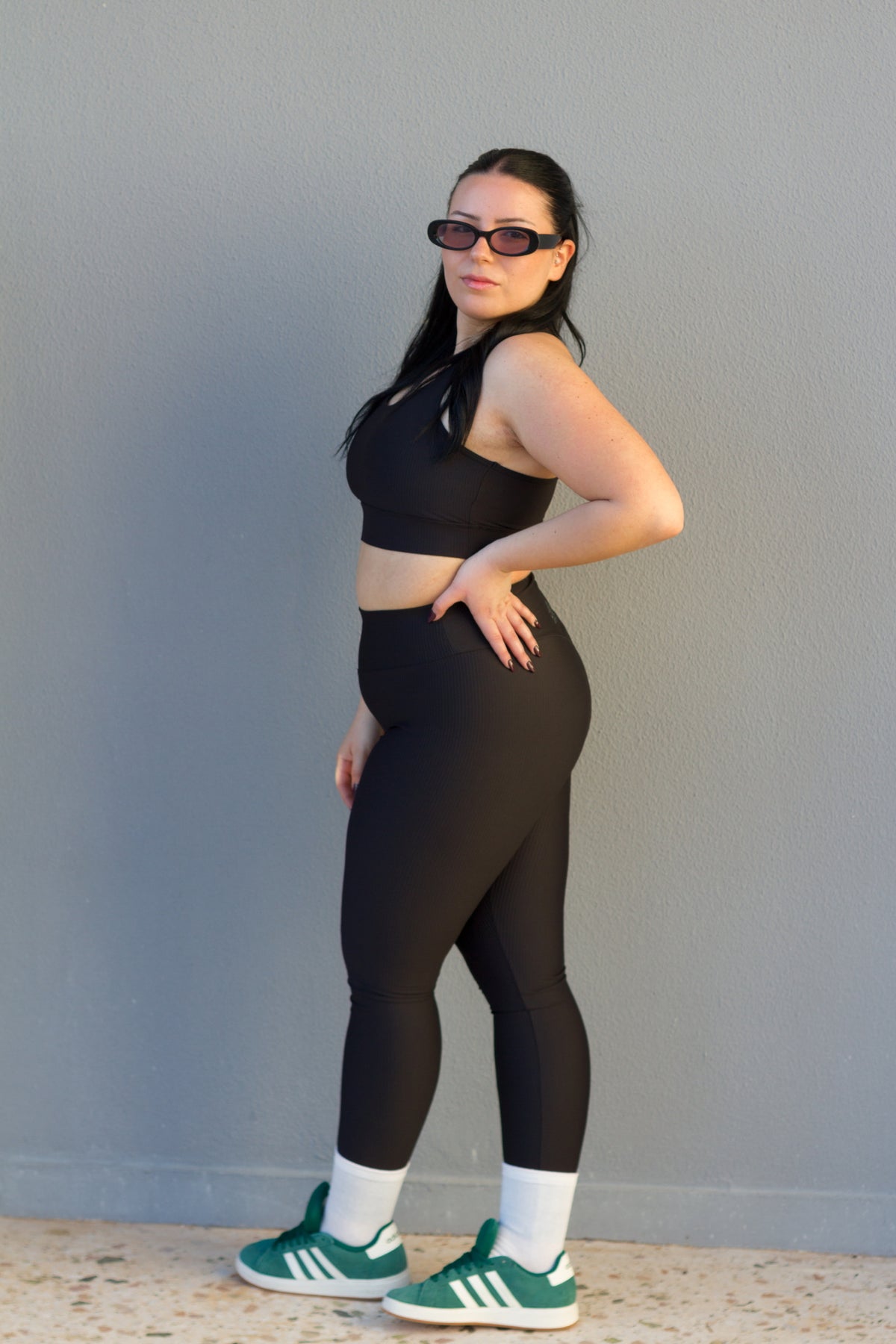 Woman wearing a black sports bra and leggings with green athletic shoes against a gray background