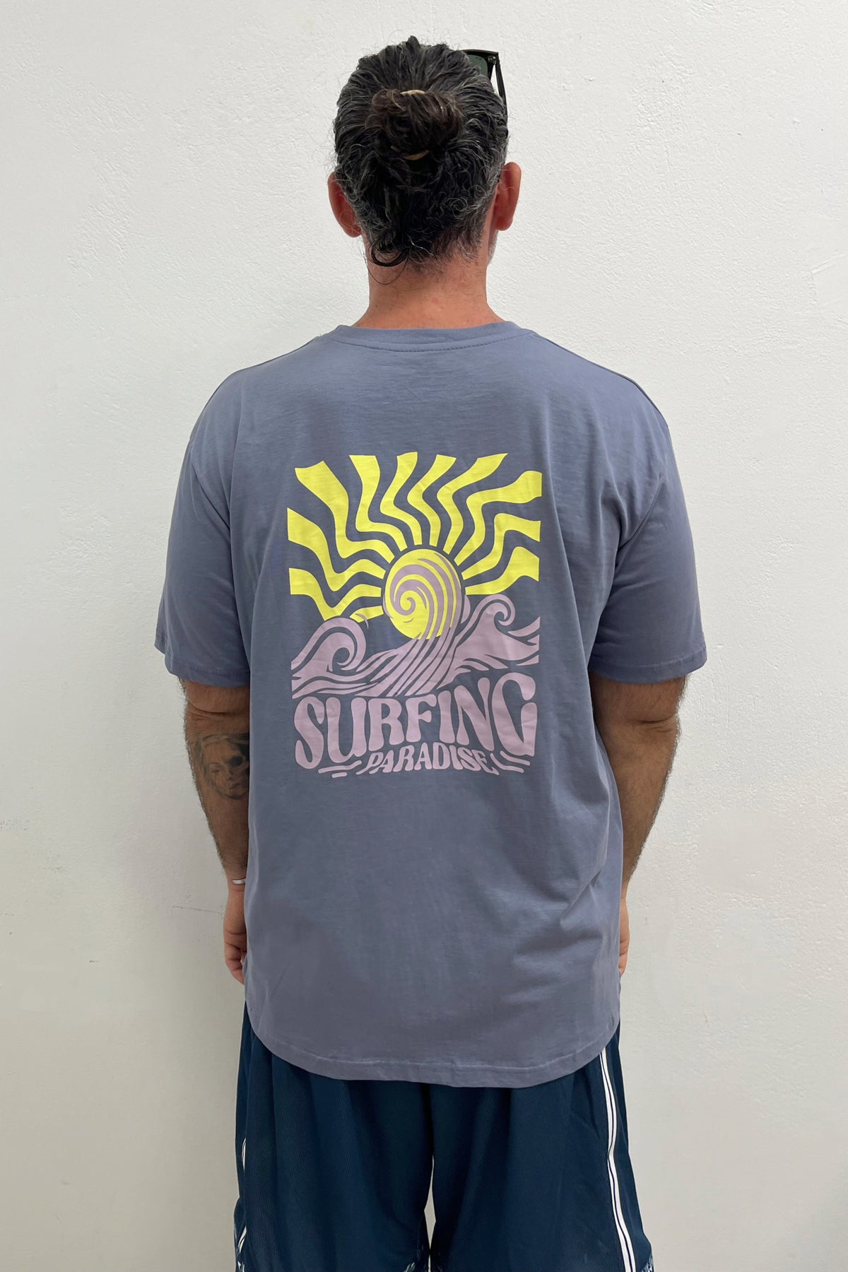 Person wearing a gray t-shirt with a yellow and purple 'Surfing Paradise' design on the back.