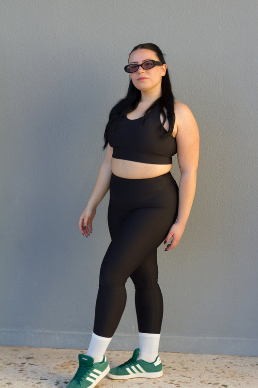 Person wearing a black sports bra and leggings set with green sneakers against a gray wall.