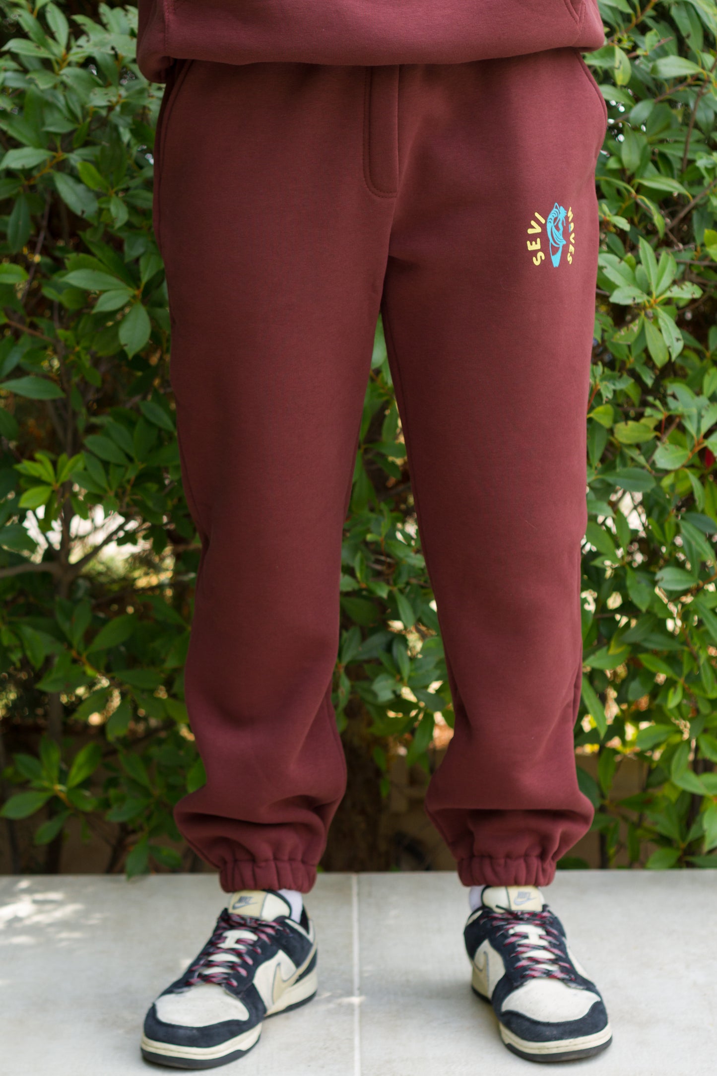 Maroon sweatpants with a colorful logo worn by a person, standing on a concrete surface with green foliage in the background.