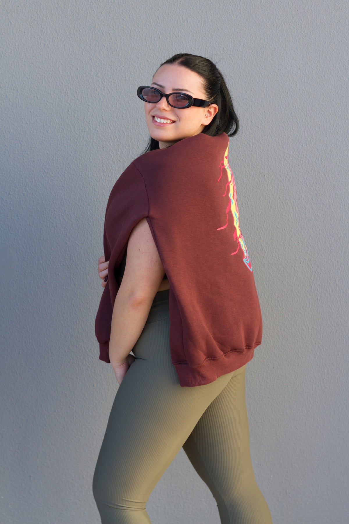 Woman wearing a maroon sweatshirt with a graphic design on the back against a plain wall.