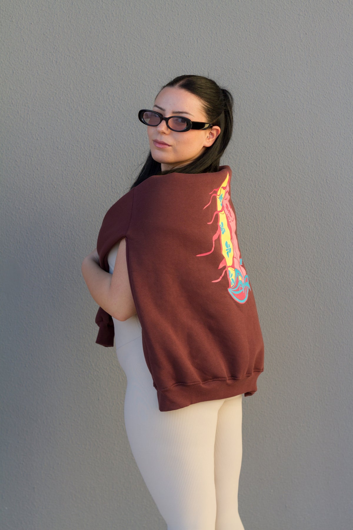 Person wearing a maroon sweatshirt with a colorful design on the back, standing against a plain wall.