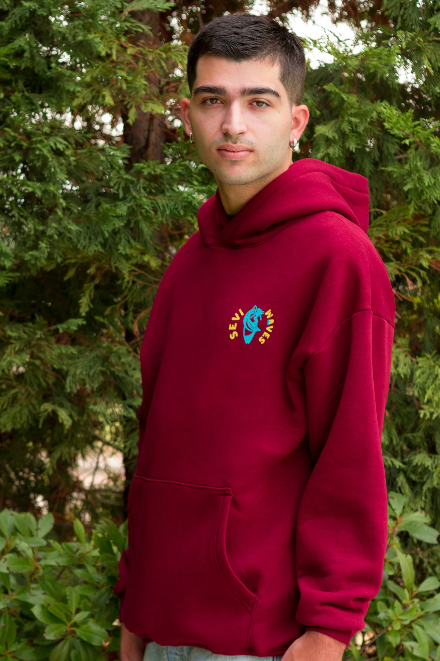 Person wearing a custom maroon hoodie with a logo in front of green foliage