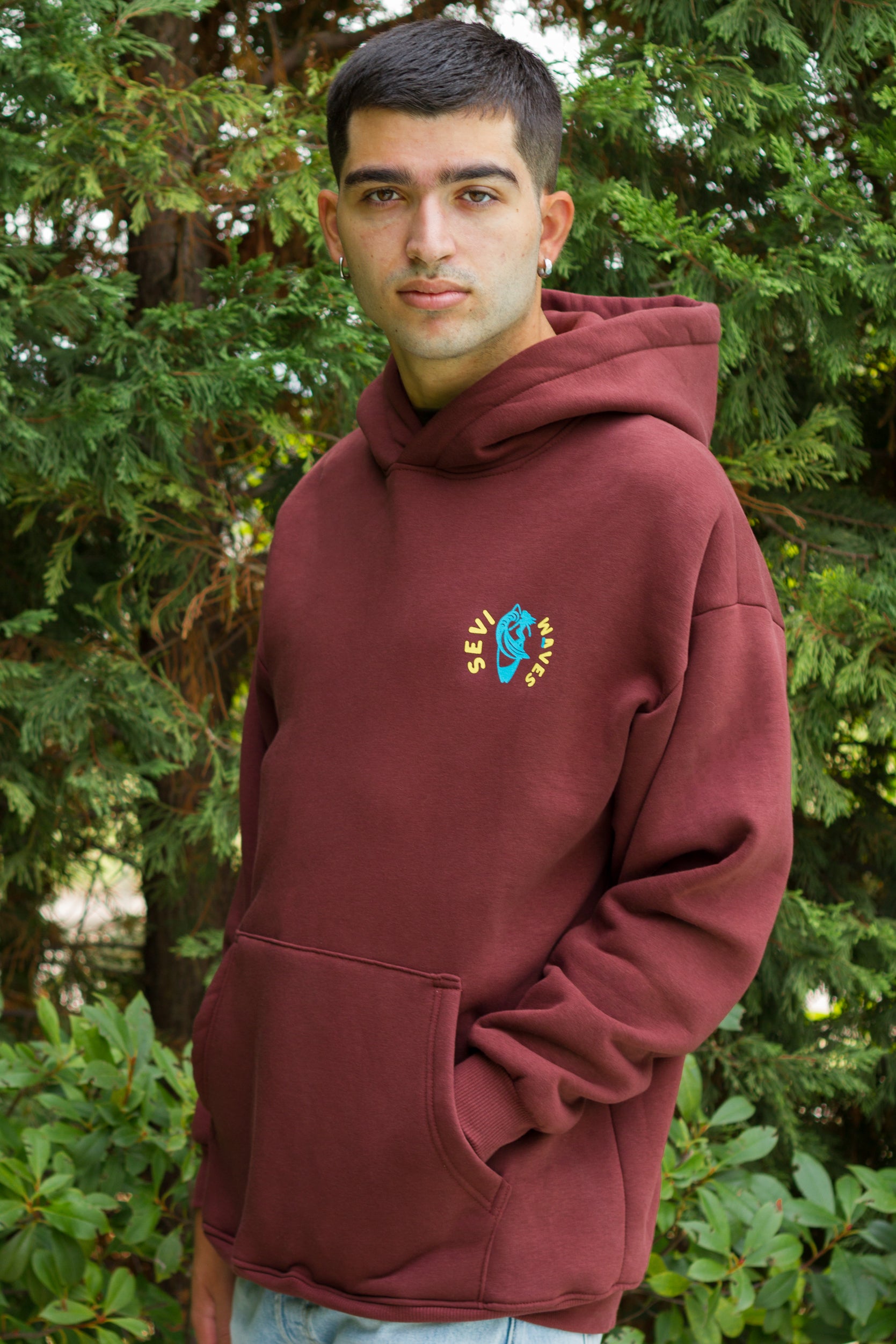 Man wearing a maroon hoodie with a custom logo in front of green foliage