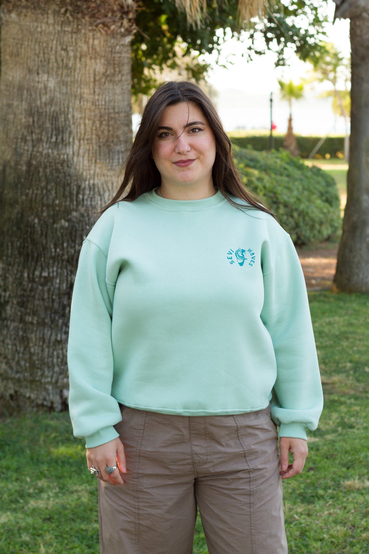 A woman standing outdoors in front of a tree wearing a light mint green crewneck sweatshirt with logo Sevi Waves on the left chest and brown pants.