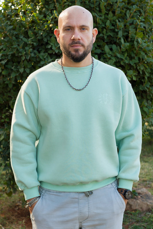 A man wearing a light mint green crewneck sweatshirt and light gray pants, standing outdoors in front of a green hedge.
