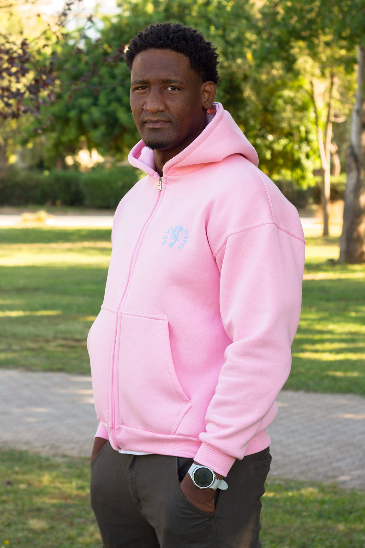 A man wearing a light pink zip-up hoodie with a small light blue circular logo on the left chest, standing outdoors in a park with green grass and trees in the background.
