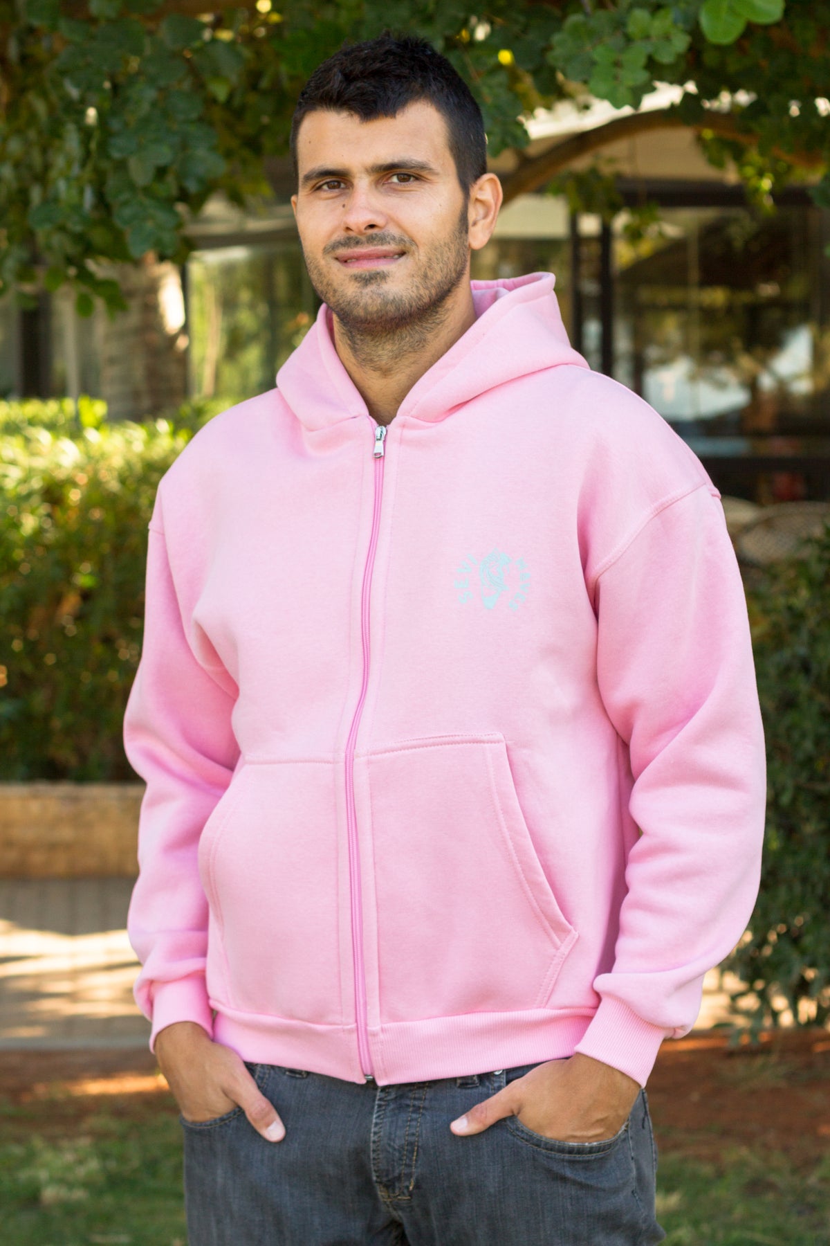 A man wearing a light pink zip-up hoodie with a small light blue circular logo on the left chest, standing outdoors in front of green foliage.