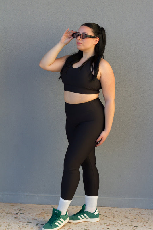 Woman wearing a black athletic outfit with green sneakers against a gray wall.