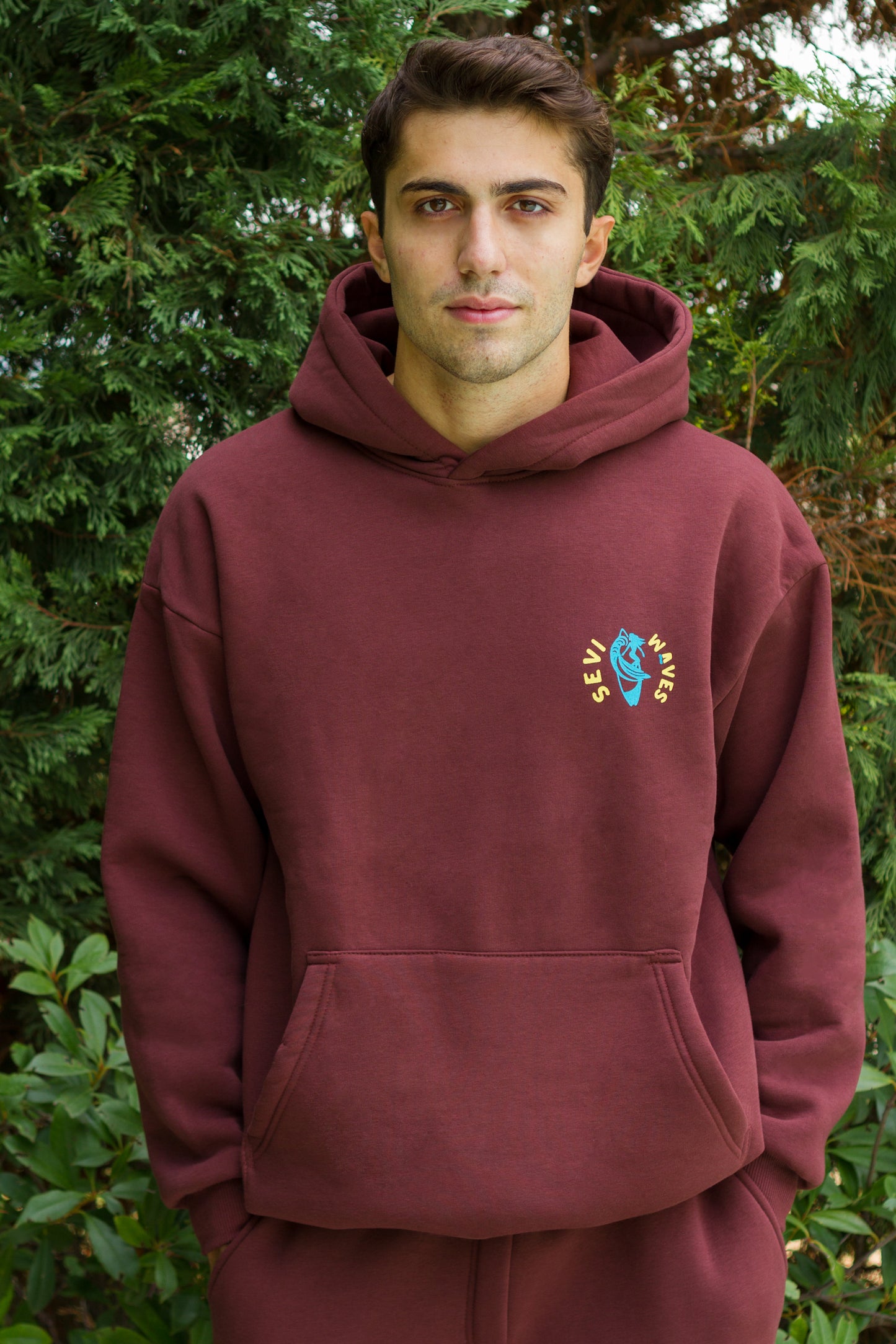 Man wearing a maroon hoodie with a colorful logo against a green foliage background