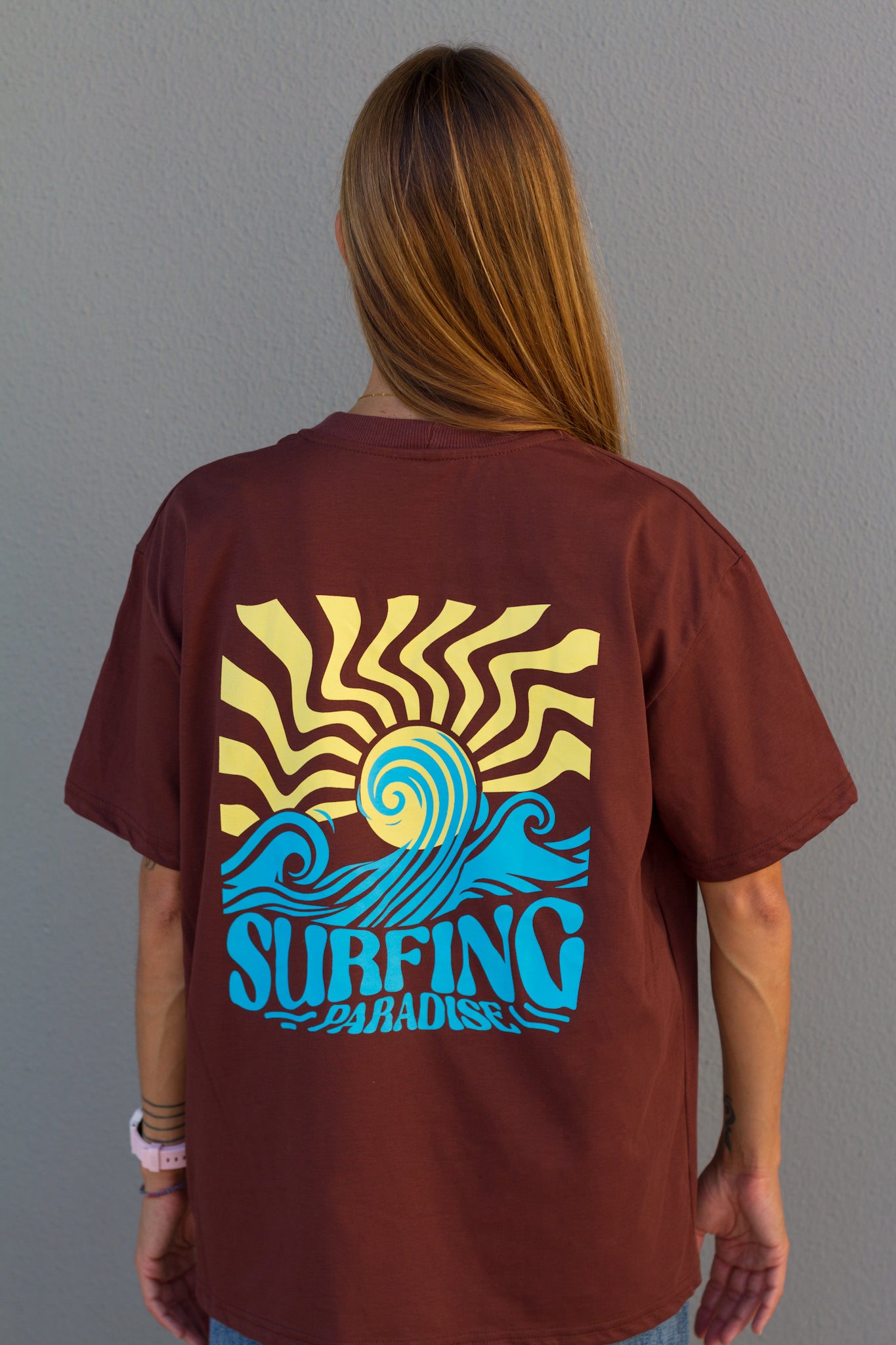 Person wearing a brown t-shirt with a 'Surfing Paradise' design on the back against a gray background