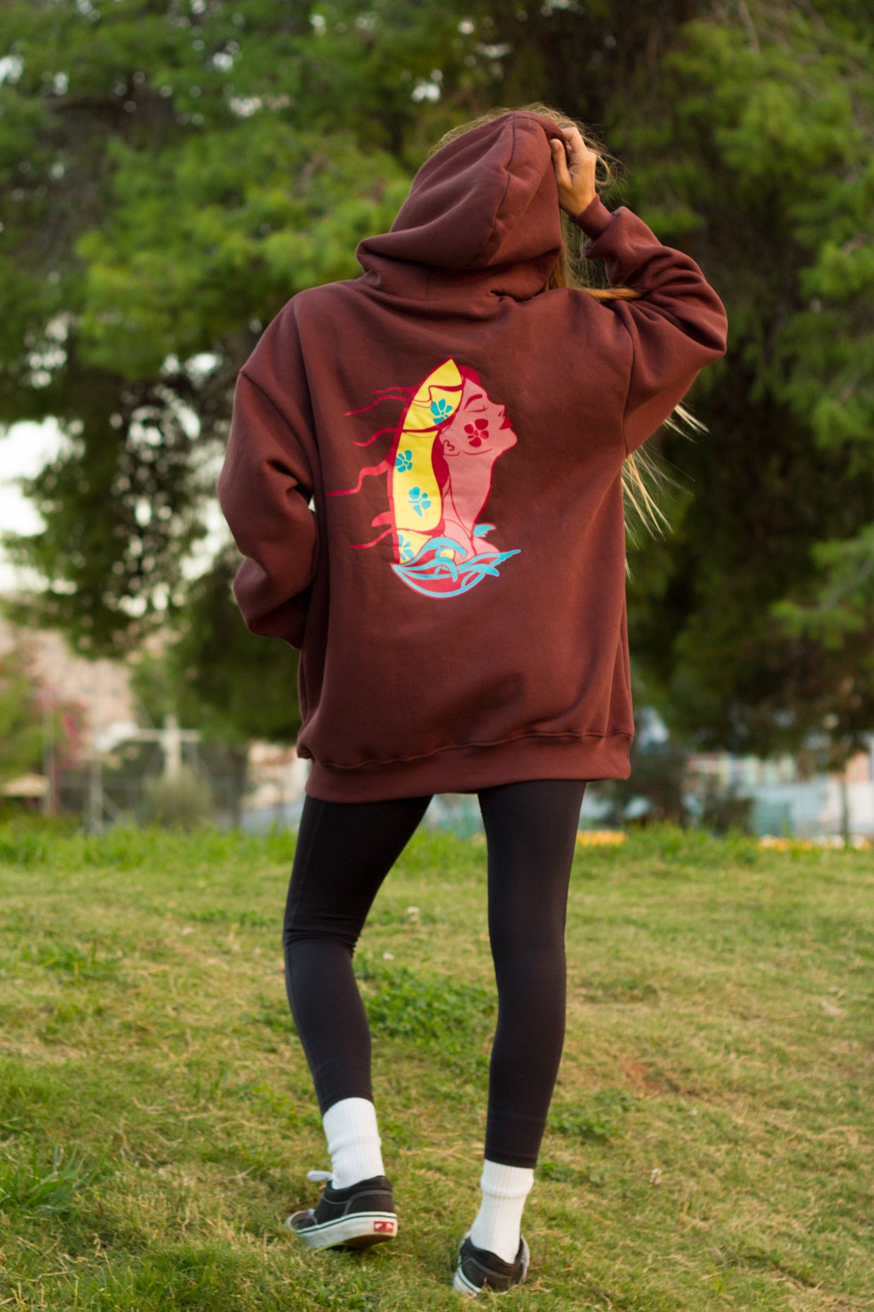 Person wearing a brown hoodie with a colorful graphic on the back, standing in a grassy area with trees in the background.