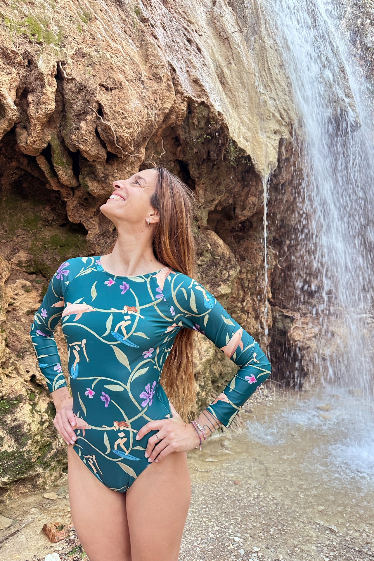 Woman in a green patterned swimsuit standing in front of a waterfall.