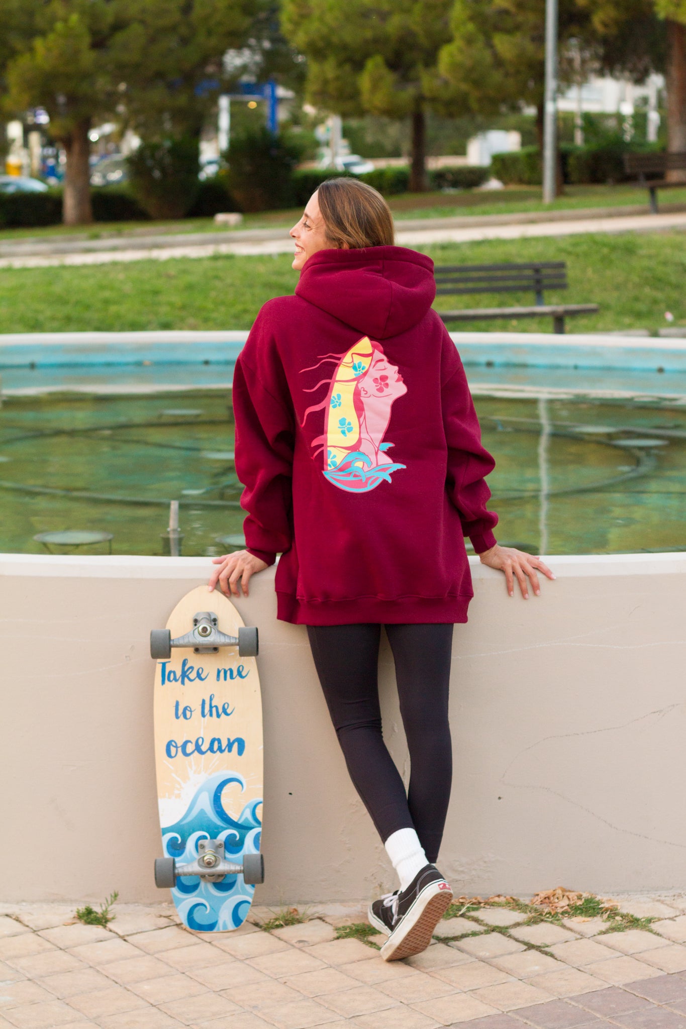 Person wearing a maroon hoodie with a colorful design, holding a skateboard with text, standing by a water feature.