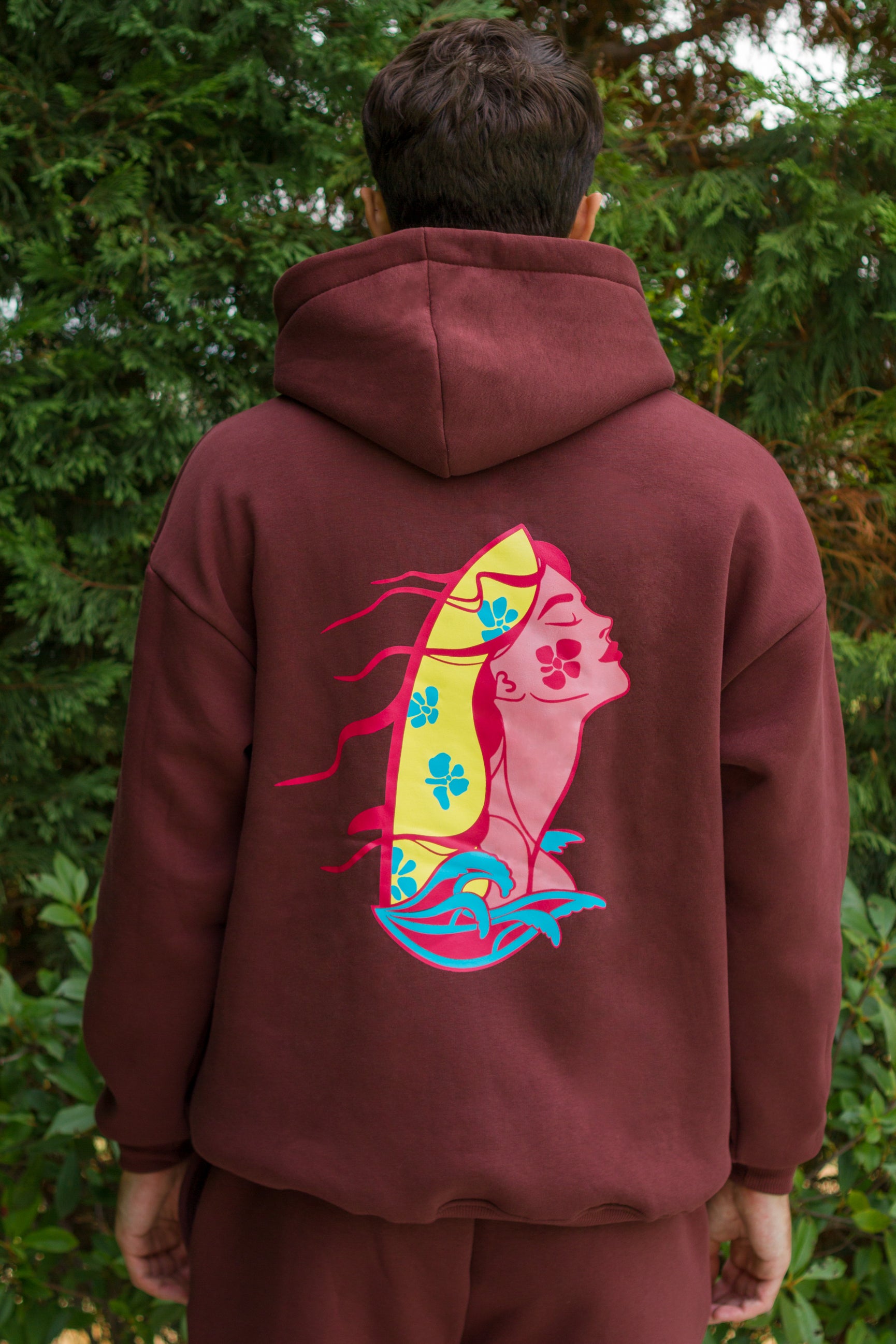 Person wearing a maroon hoodie with a colorful custom graphic on the back, standing against a green leafy background.