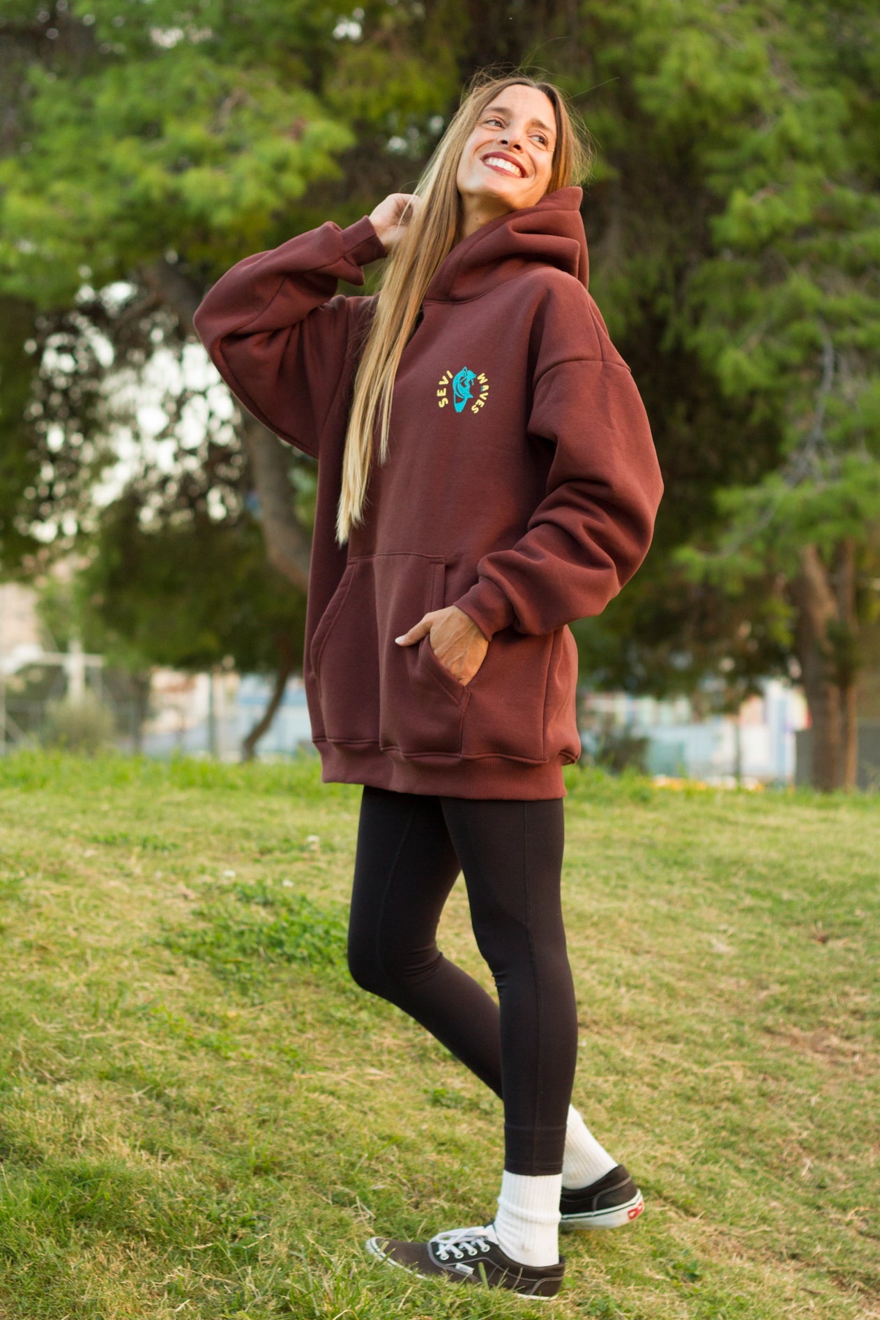 Person wearing a maroon hoodie with a logo, standing outdoors on grass with trees in the background