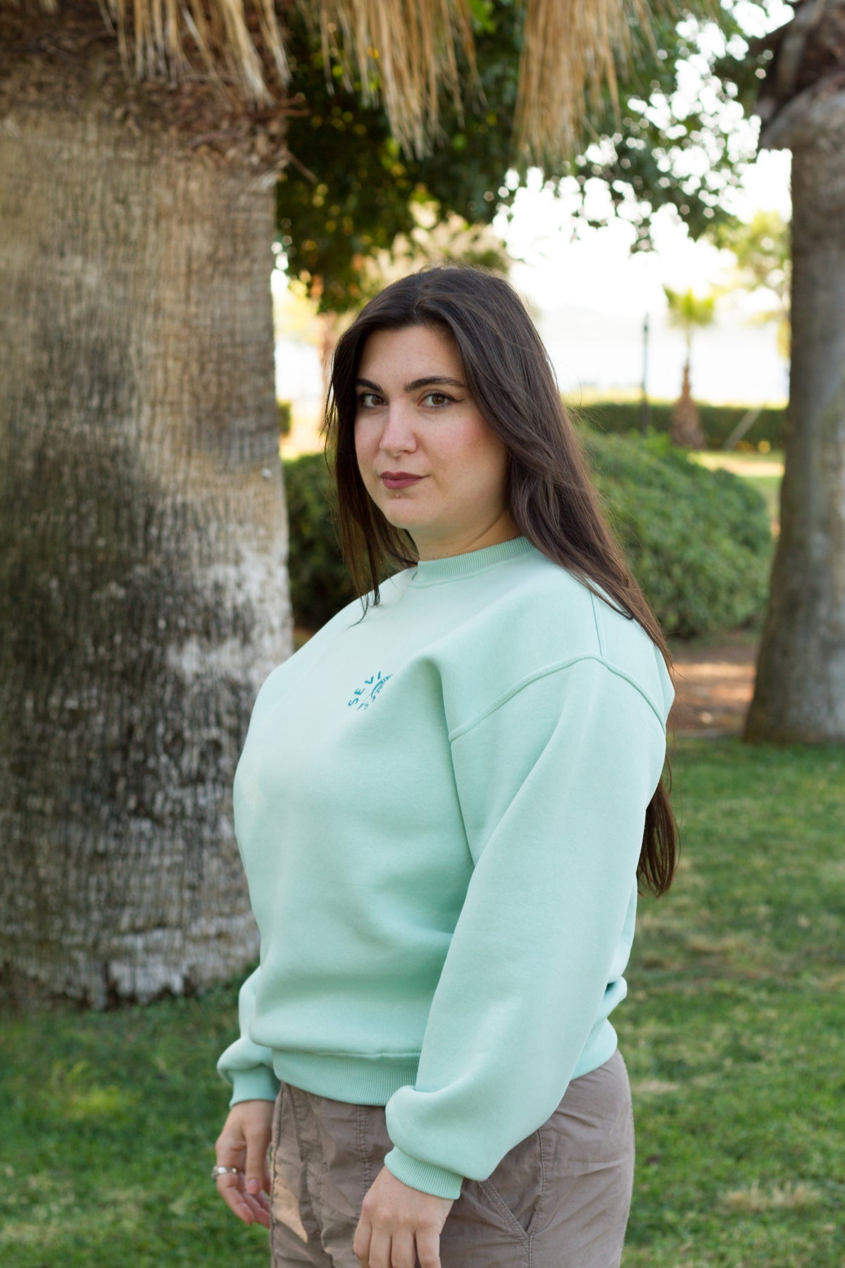 A woman wearing a mint green crewneck sweatshirt and brown pants, standing outdoors in front of a large palm tree trunk.