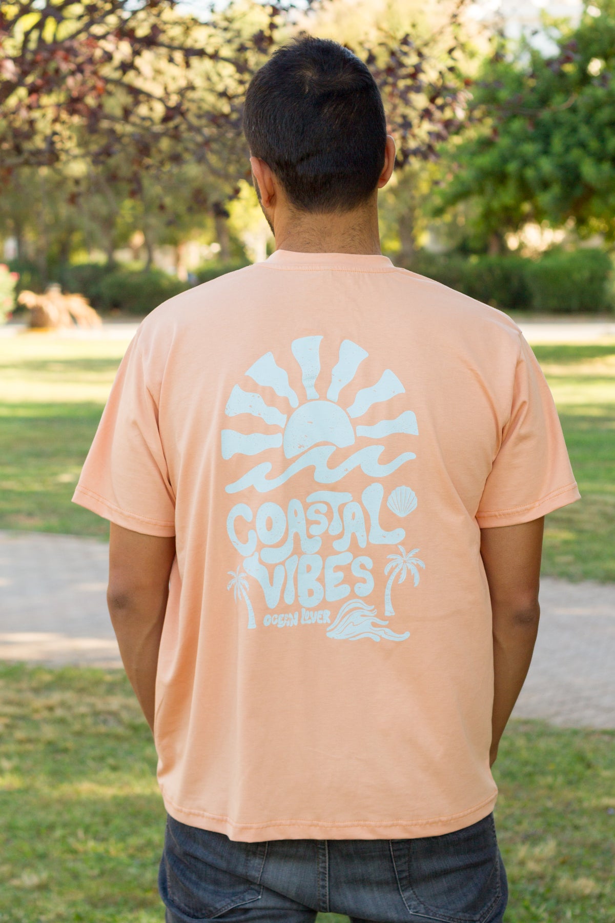 A man viewed from behind wearing a peach-colored t-shirt with a large white graphic on the back that reads 'COASTAL VIBES' with a sun and palm tree design, set in an outdoor park environment by Sevi Waves 