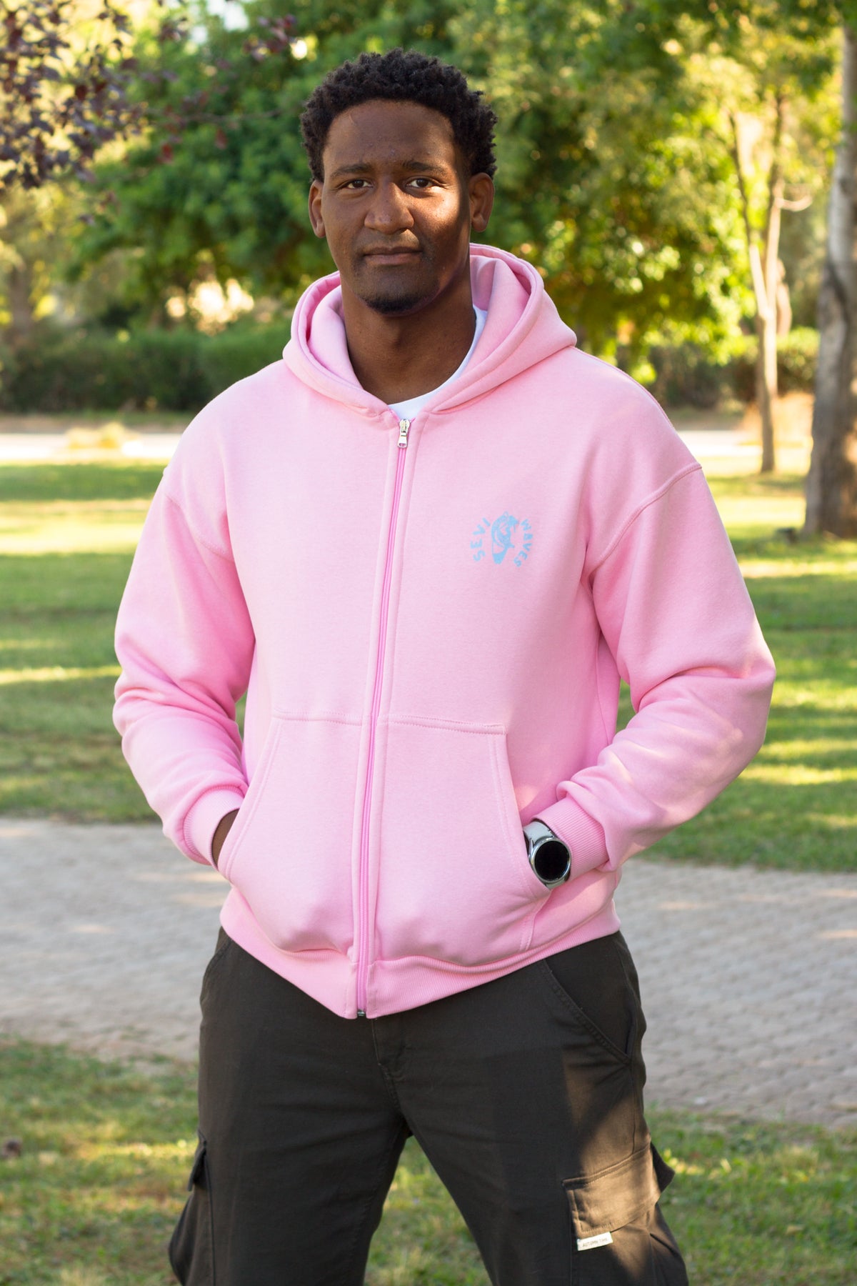A man wearing a light pink zip-up hoodie with a small light blue circular logo on the left chest, standing outdoors in a park with trees and grass in the background by Sevi Waves 