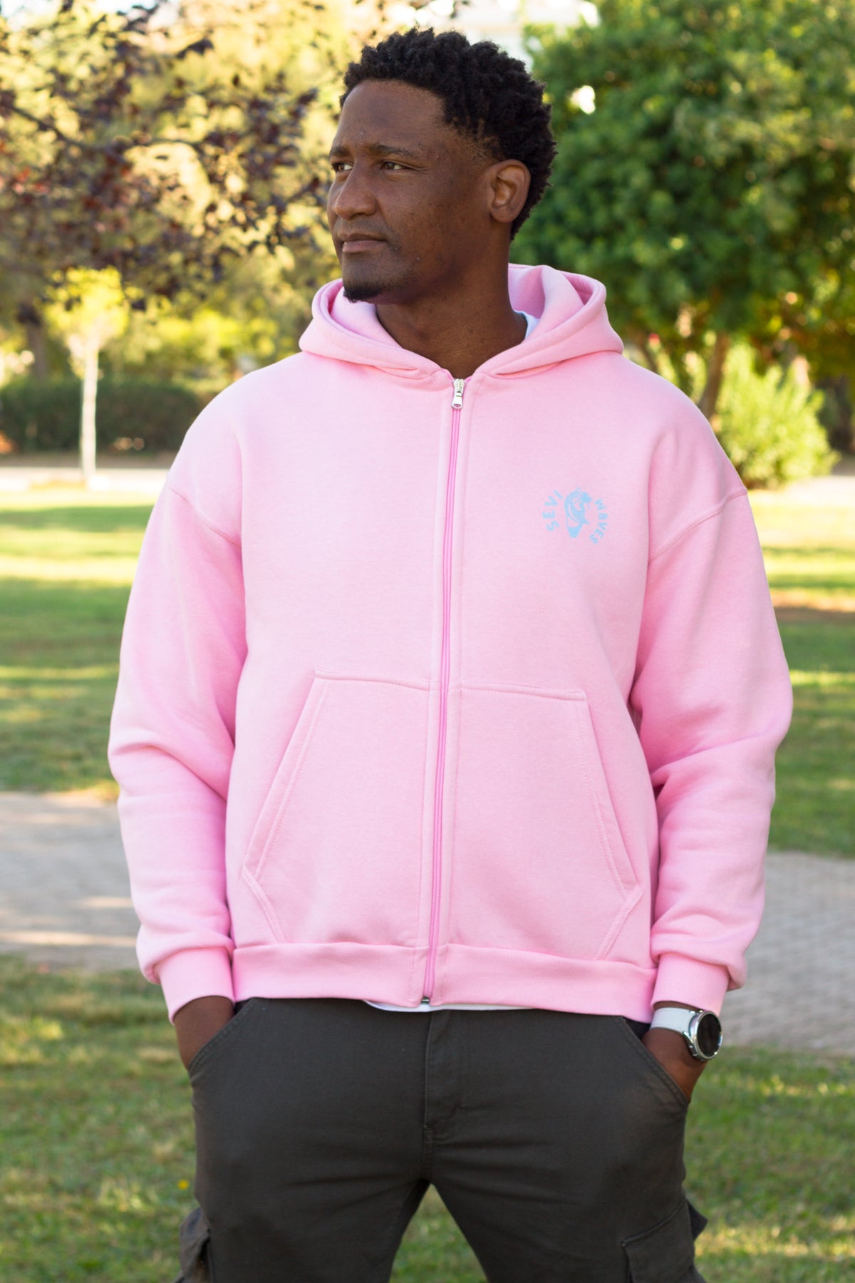 A man wearing a light pink zip-up hoodie with a small light blue Sevi Waves logo  on the left chest, standing outdoors in a park with trees and grass in the background.