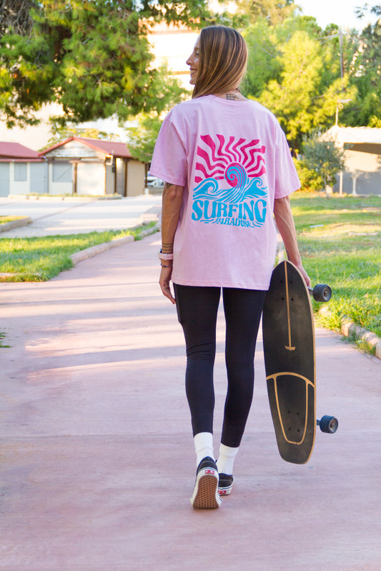 Person walking with a skateboard wearing a pink t-shirt with a 'Surfing' design.