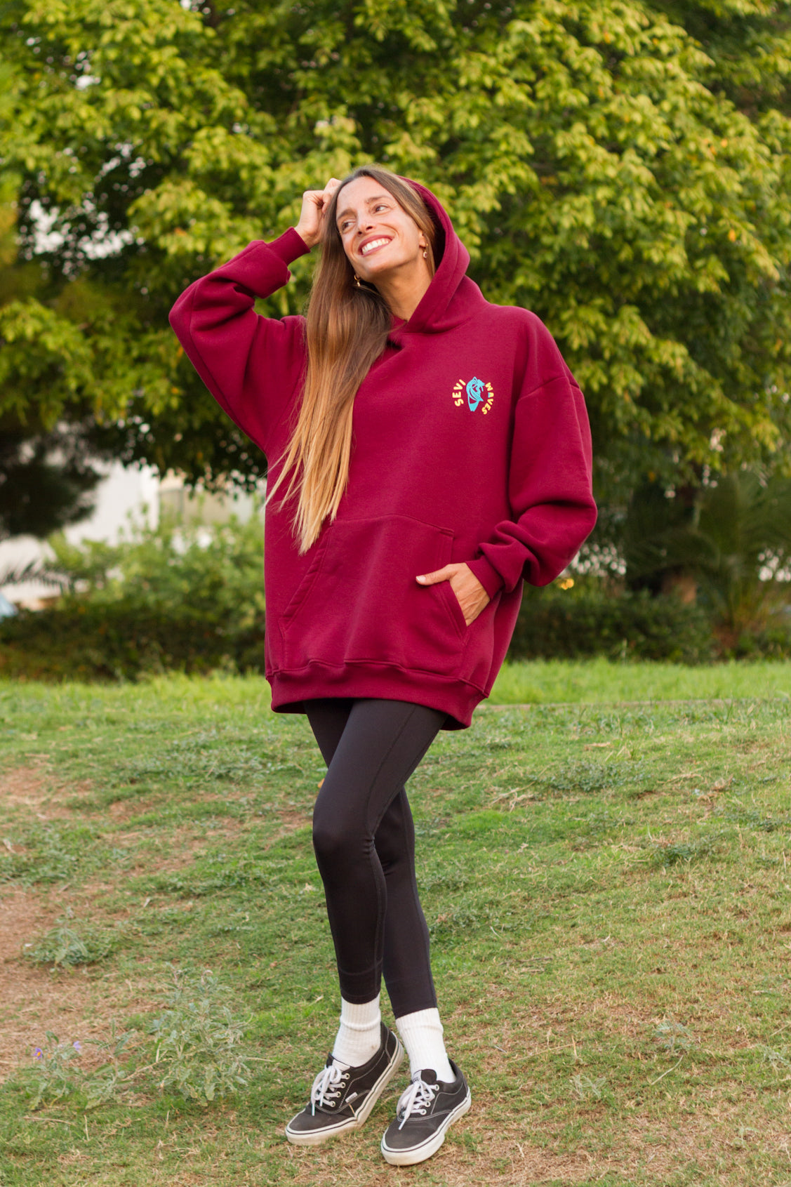 Person wearing a maroon hoodie with a logo, standing outdoors in a park.