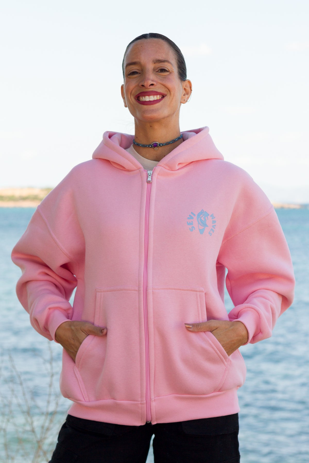 A smiling woman wearing a pink zip-up hoodie with a light blue circular logo Sevi Waves on the left chest, standing outdoors with water and a pale sky in the background.