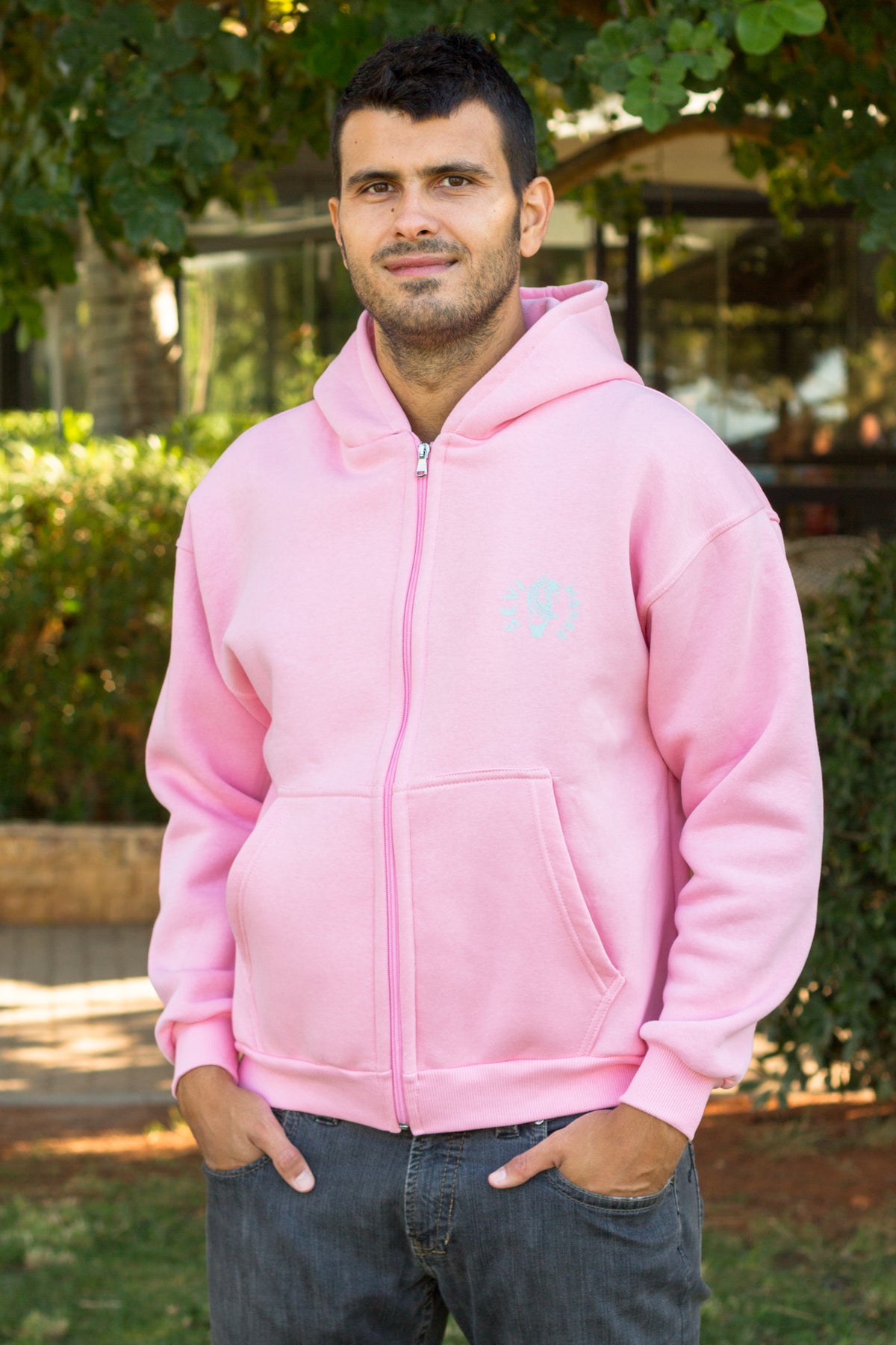 A man wearing a pink zip-up hoodie with a small light blue embroidered logo on the left chest, standing outdoors in front of green foliage.