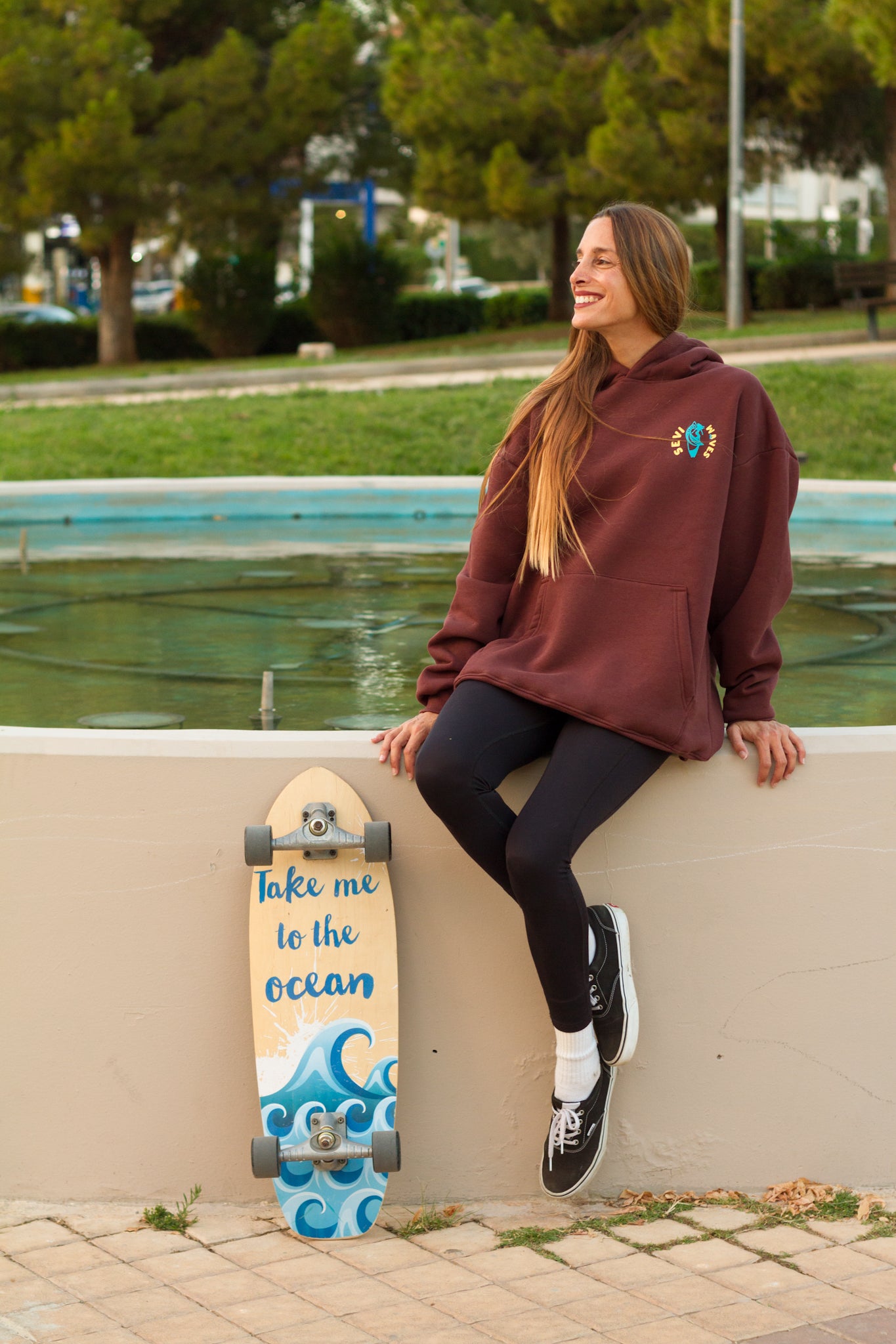 Woman sitting on a wall with a skateboard next to her, near a fountain with greenery in the background.