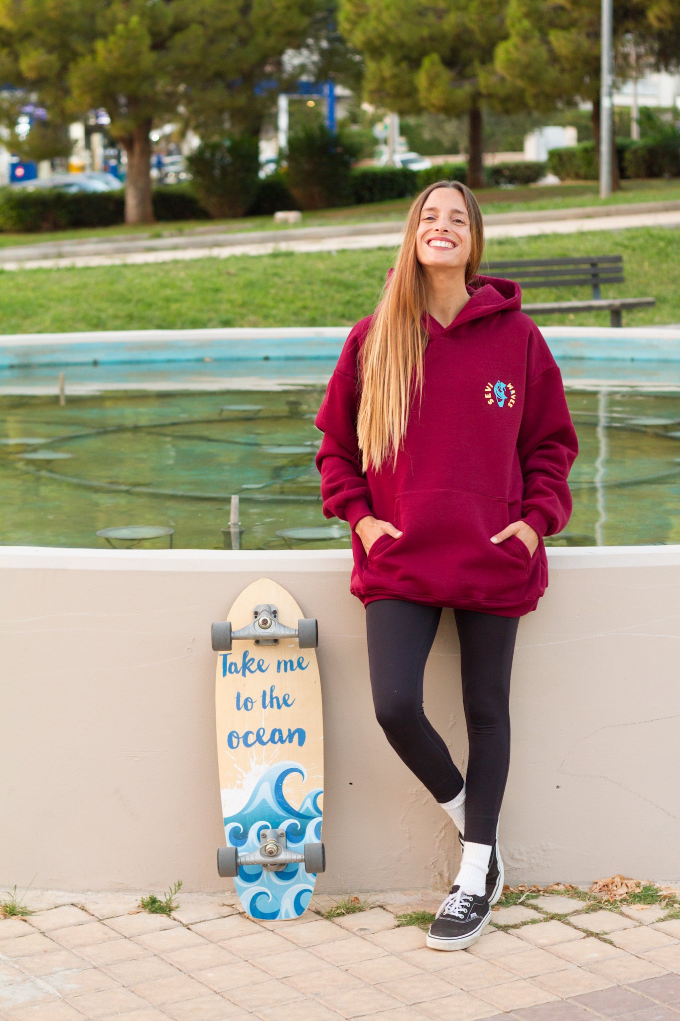 Person wearing a maroon hoodie with a logo, standing next to a skateboard with ocean design, in an outdoor setting.