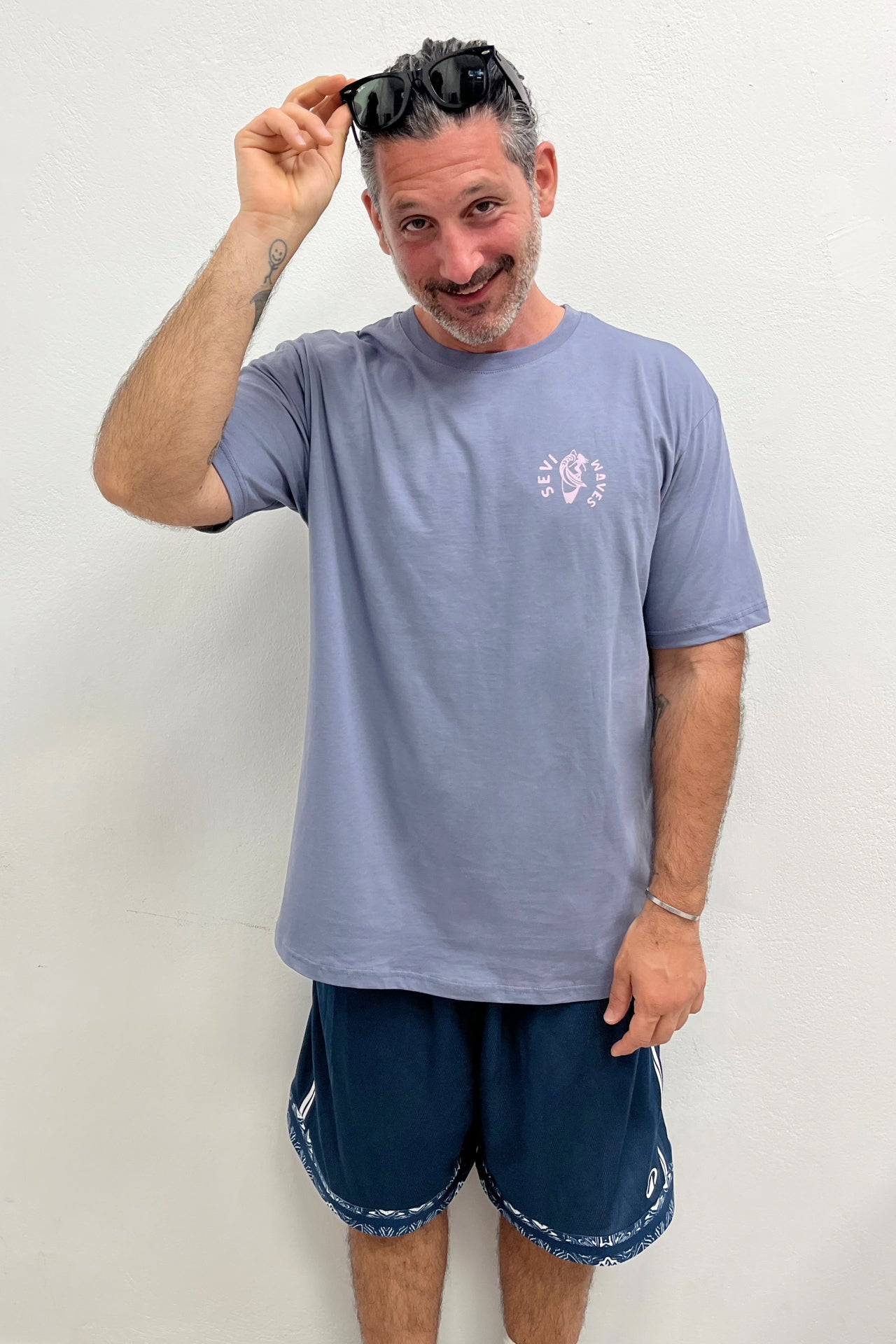 Man wearing a light blue t-shirt and dark shorts, holding sunglasses above his head against a plain background.