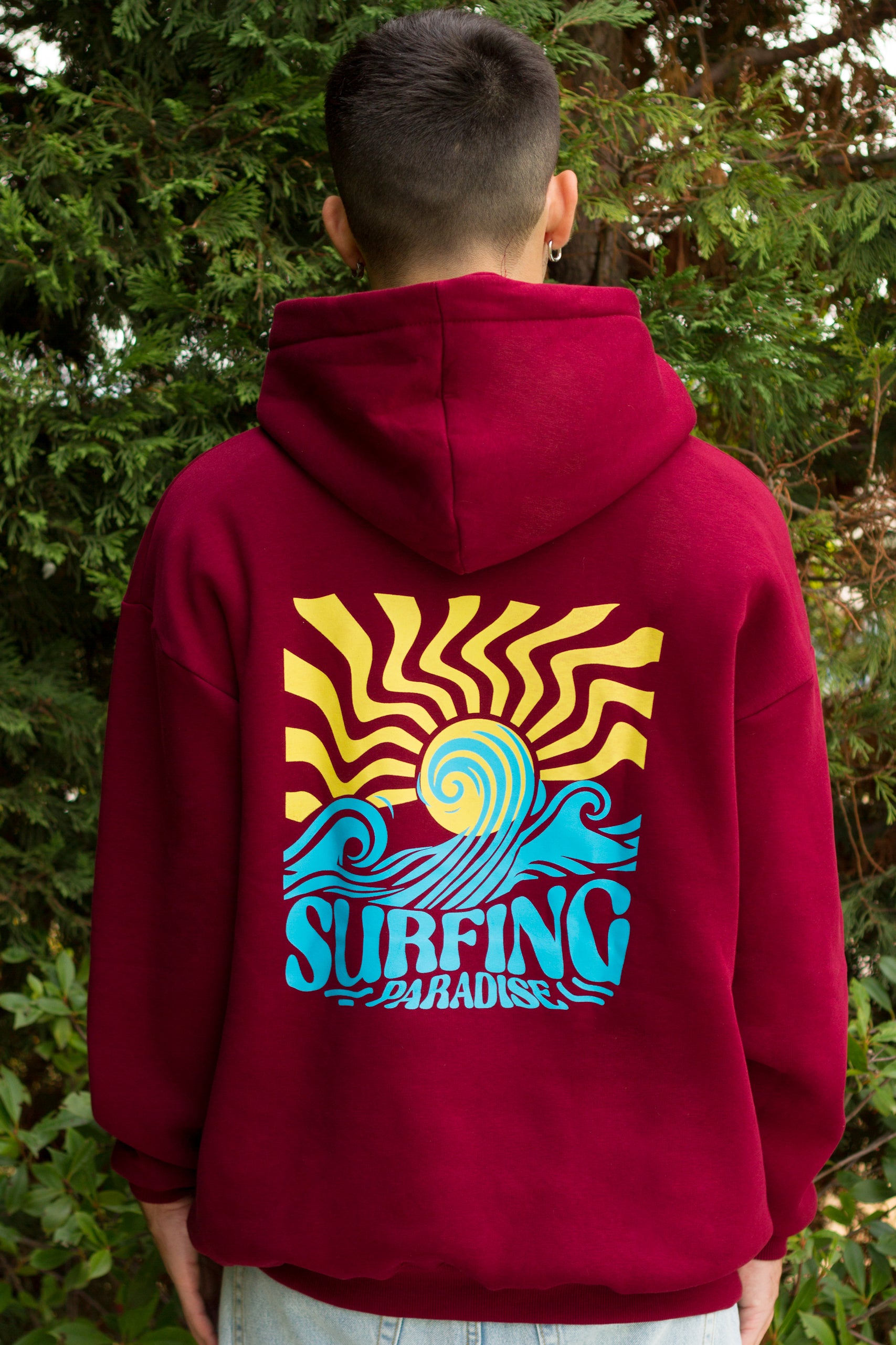 Person wearing a red hoodie with a colorful 'Surfing Paradise' design on the back, standing in front of green foliage.