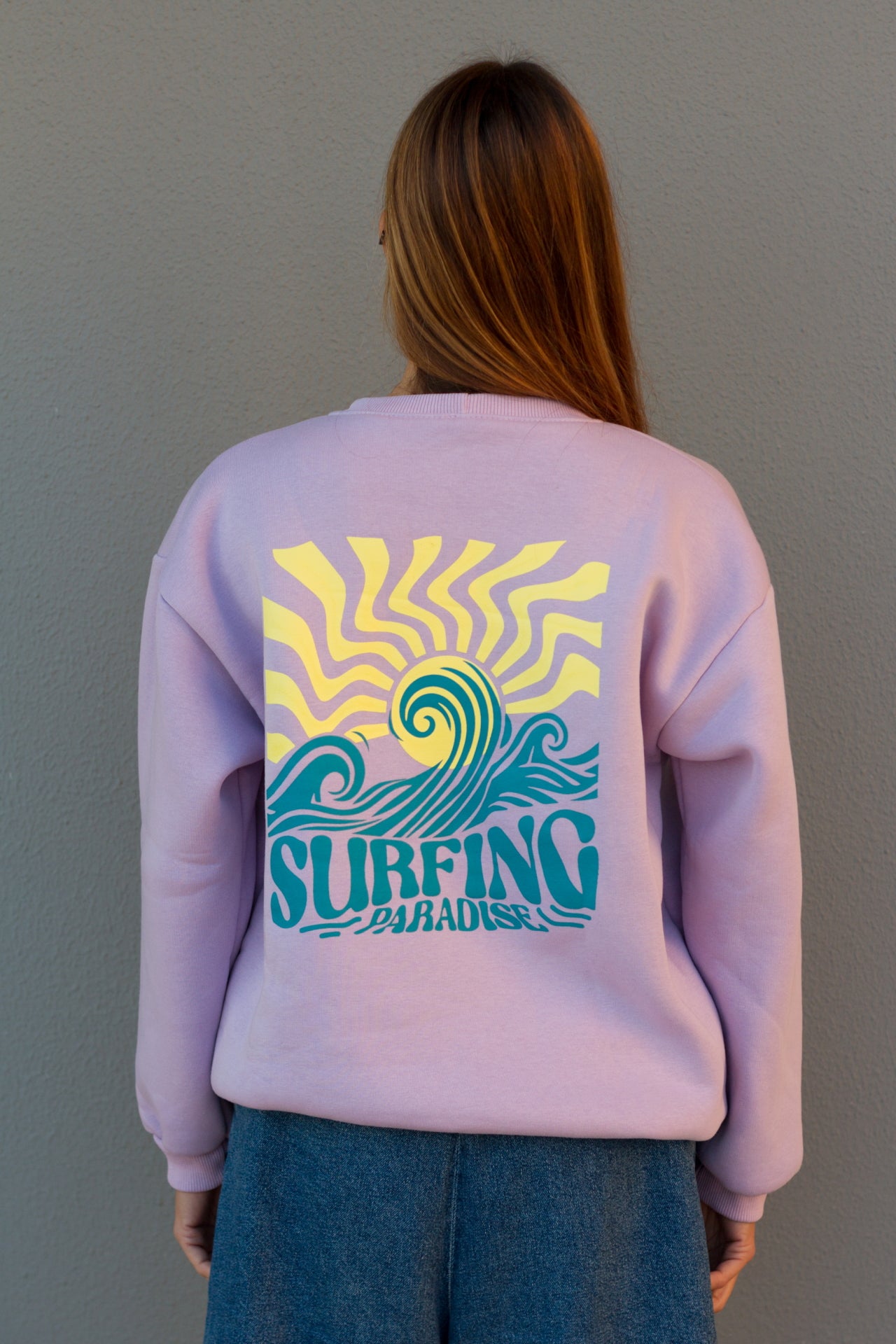 Person wearing a light purple sweatshirt with a colorful 'Surfing' graphic on the back against a gray background