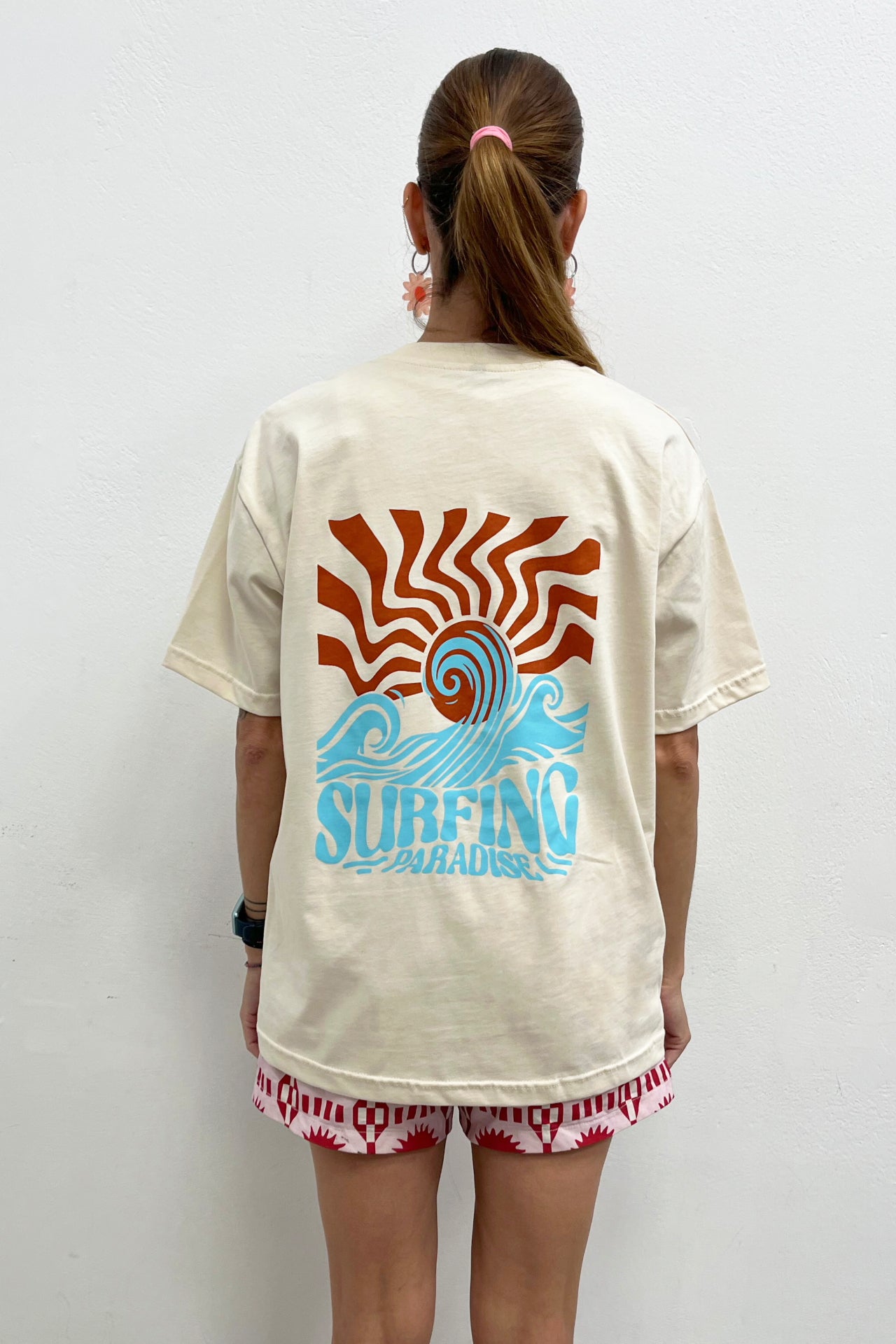 Person wearing a beige t-shirt with a colorful 'Surfing' design on the back, standing against a white background.