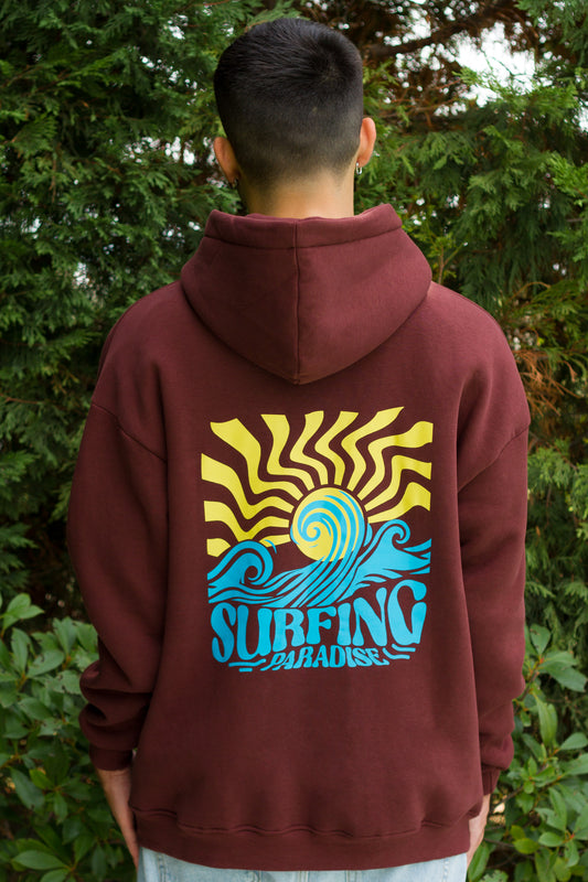 Person wearing a maroon hoodie with a 'Surfing Paradise' design on the back, standing in front of green foliage.