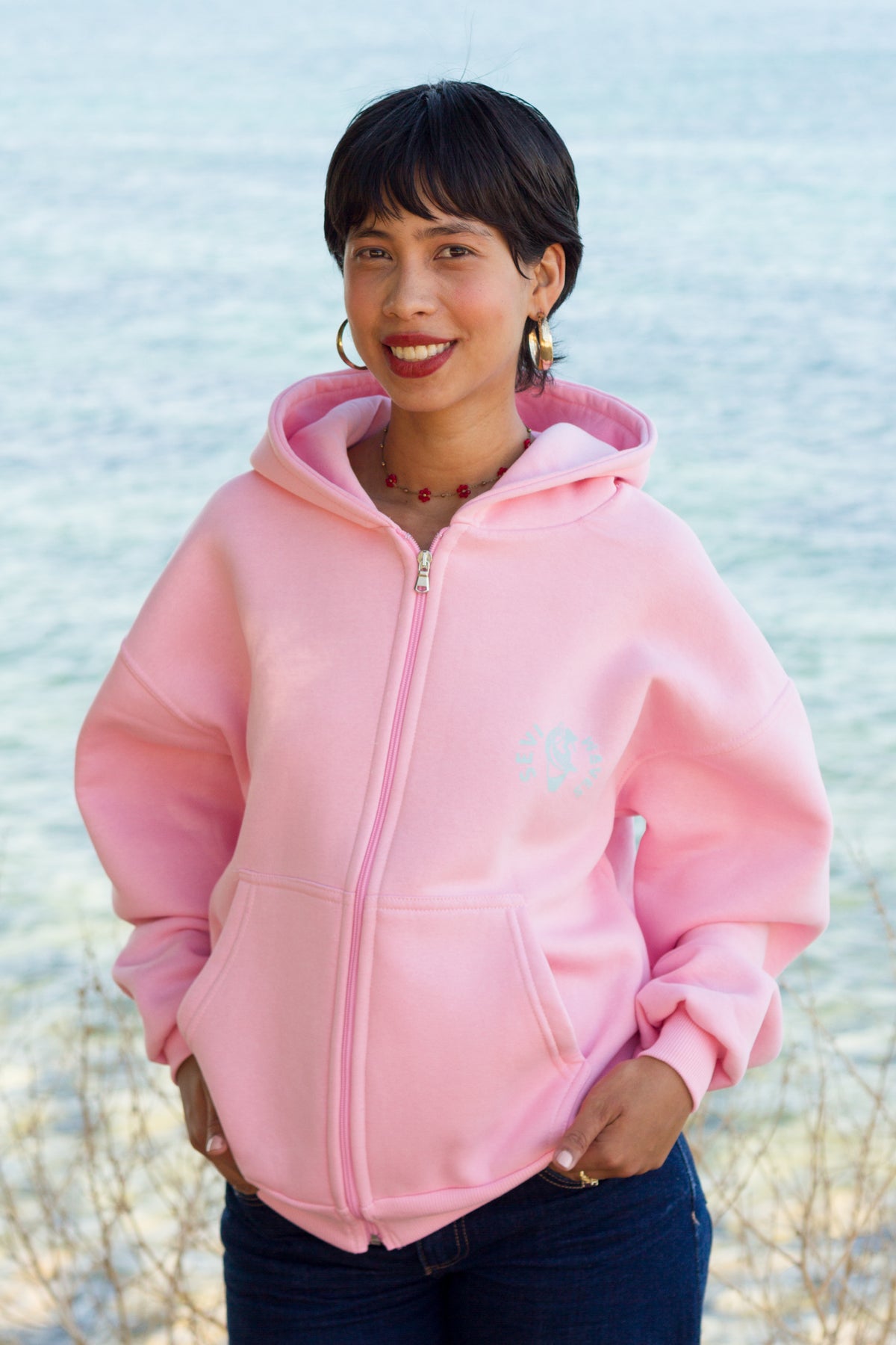 A woman wearing a light pink zip-up hoodie with a small light blue graphic on the left chest, standing outdoors with a blurred body of water in the background.