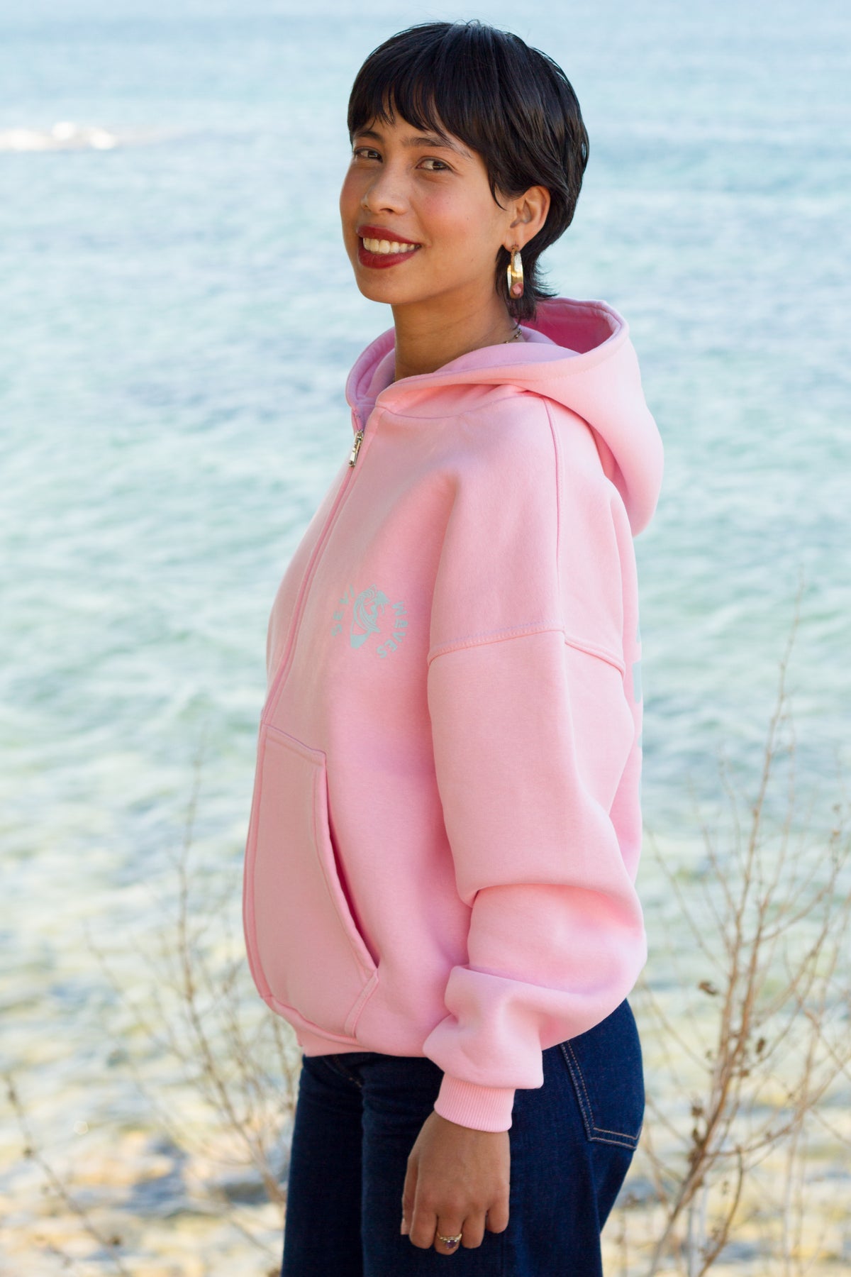 A woman wearing a light pink pullover hoodie with a small light blue embroidered graphic on the left chest, standing outdoors near the water by Sevi Waves new collection 