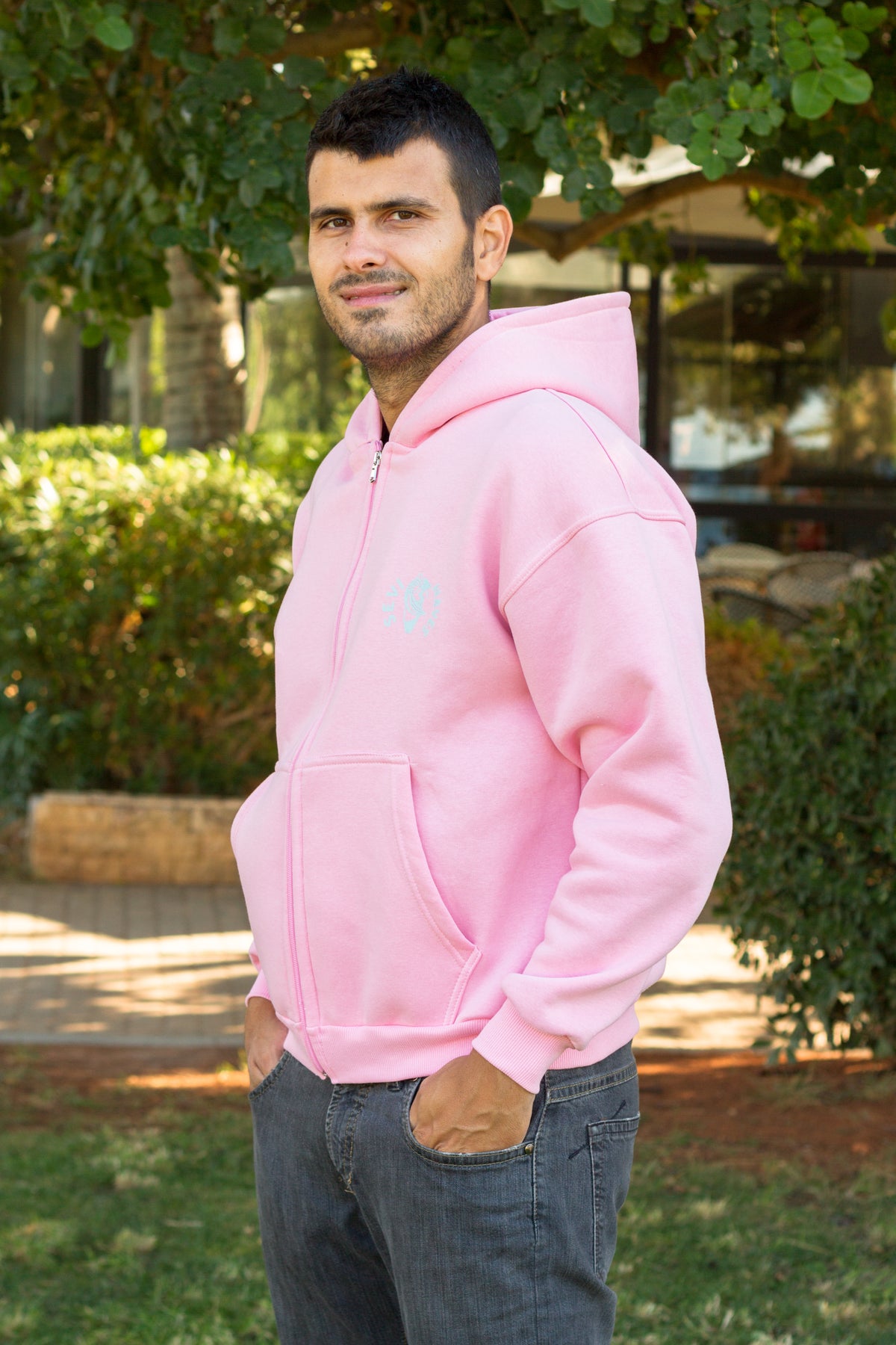 A man wearing a light pink zip-up hoodie with a small light blue graphic on the left chest Sevi Waves , paired with dark gray jeans, standing outdoors in a park-like setting with green trees and bushes.