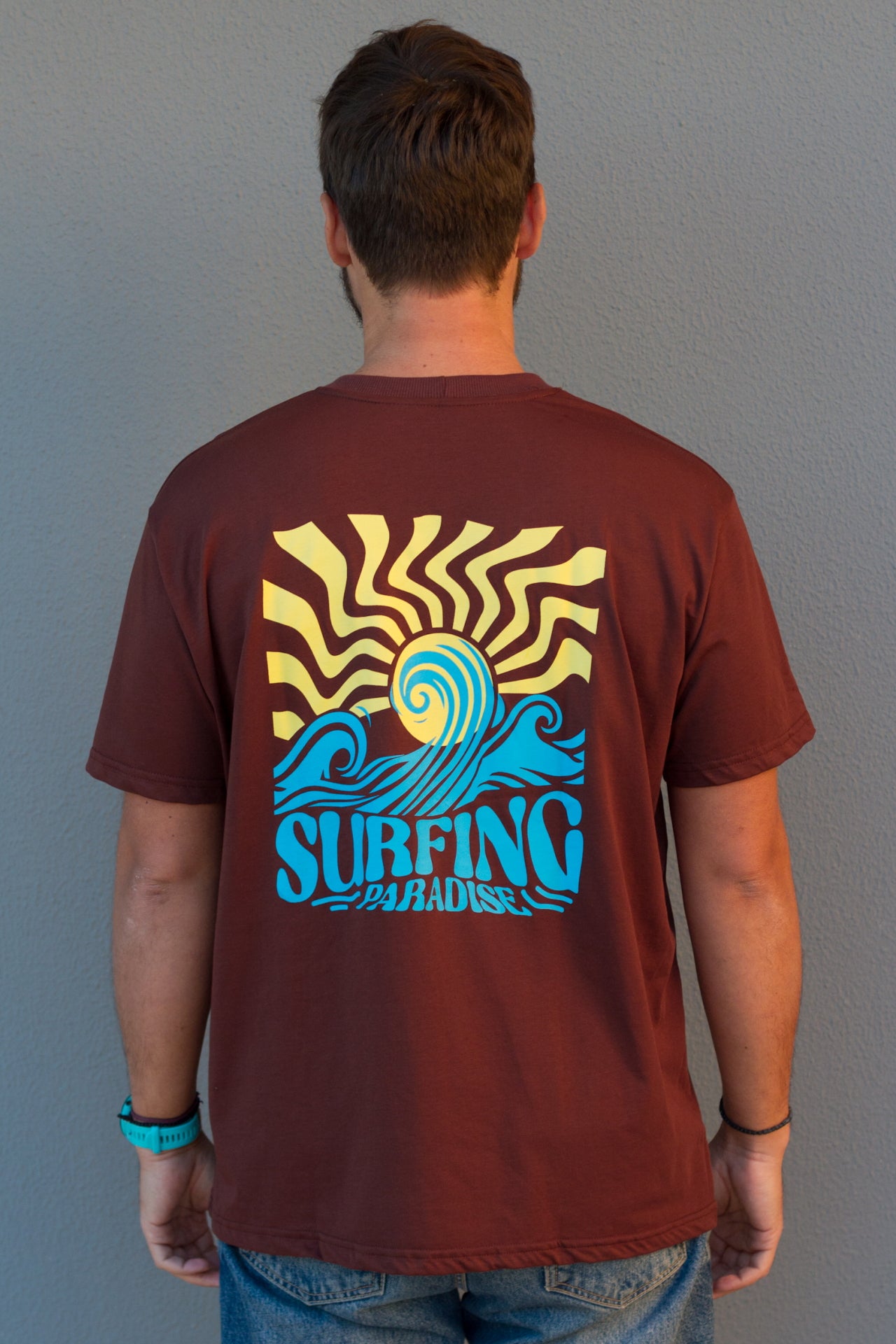 Man wearing a maroon t-shirt with a 'Surfing Paradise' graphic design on the back against a gray background.