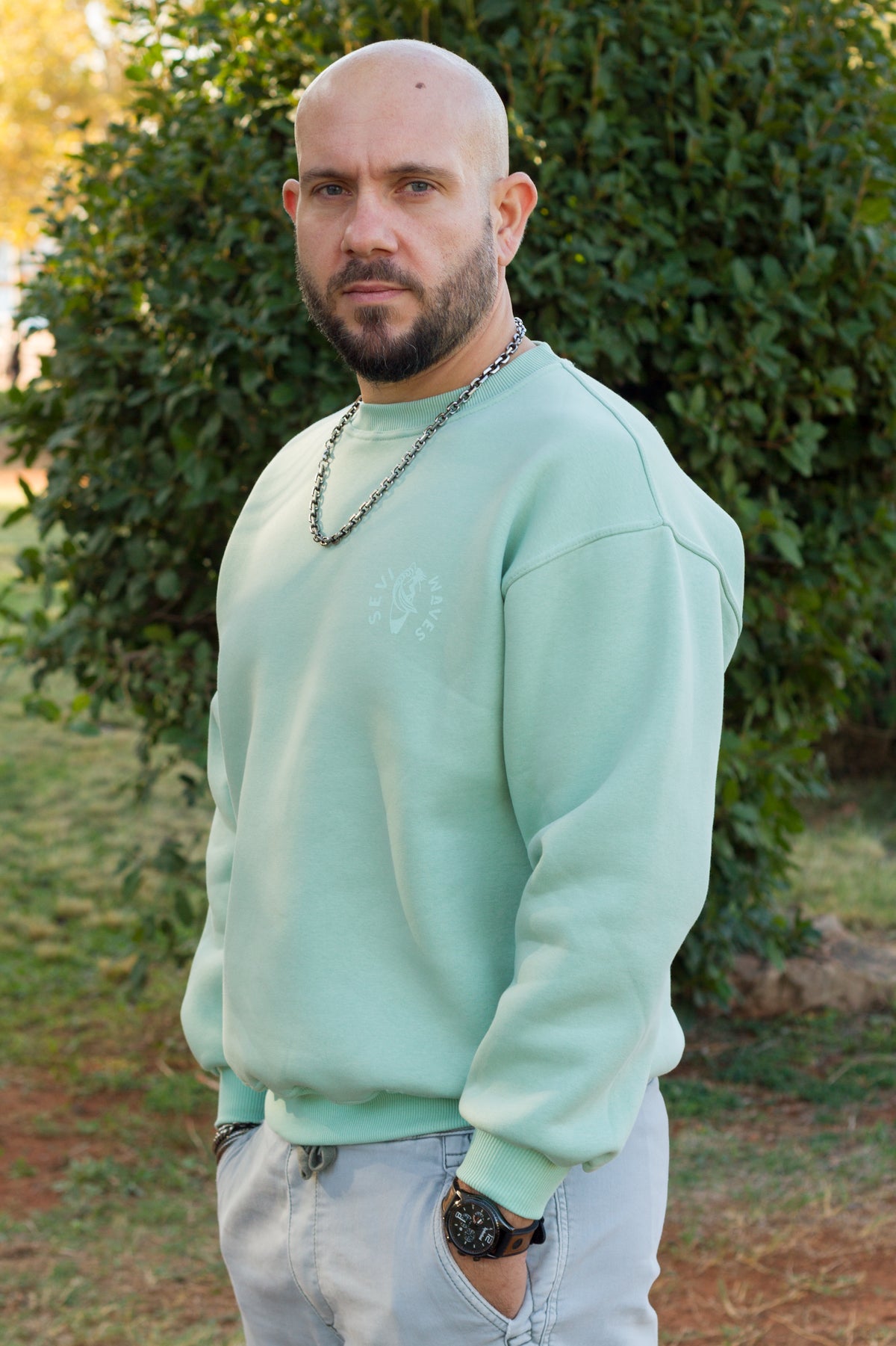 A man wearing a light mint green crewneck sweatshirt and light gray jeans, standing outdoors in front of a green hedge from Sevi Waves new collection 
